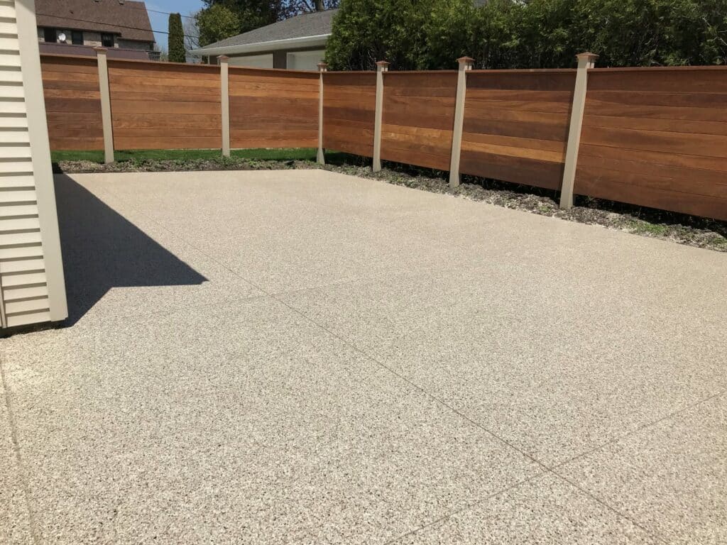 This image shows a clean and spacious concrete driveway next to a house, bordered by a tall, modern wooden fence under a clear sky.