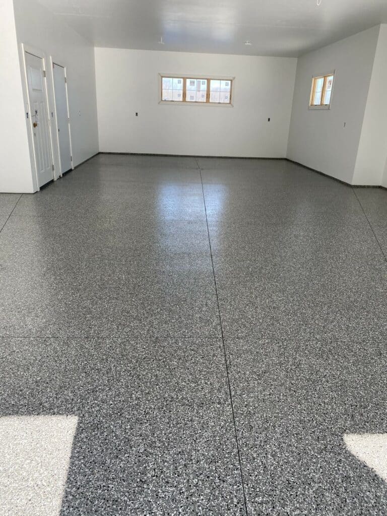 An empty room with white walls and a speckled gray epoxy floor features multiple windows, a doorway, and reflects natural light.