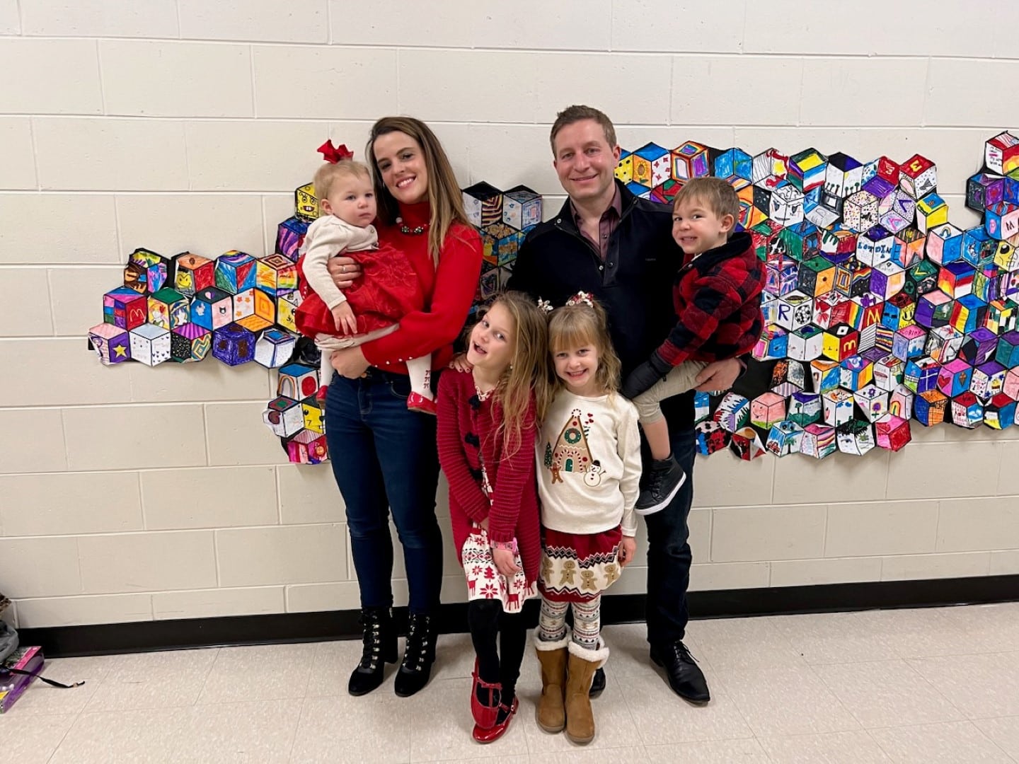 A family of five, with two adults and three children, stand smiling against a wall decorated with colorful artwork, dressed in festive attire.