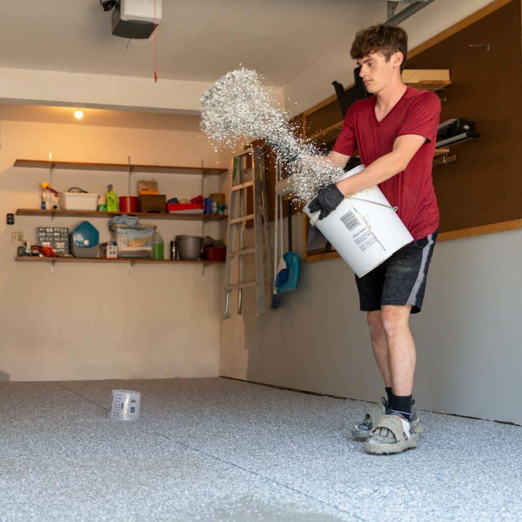 A person is pouring a substance from a white bucket onto a garage floor, creating a speckled effect. Shelves with various items line the back wall.