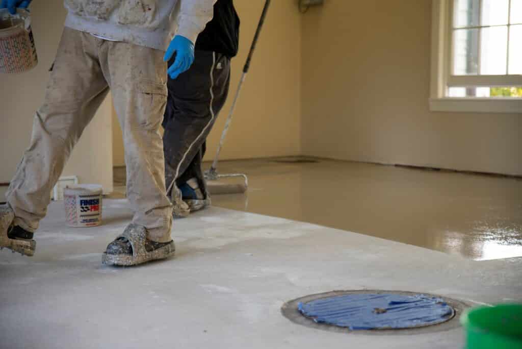 Two people are applying a light-colored epoxy or paint on a concrete floor inside a room with a window, wearing protective workwear and gloves.